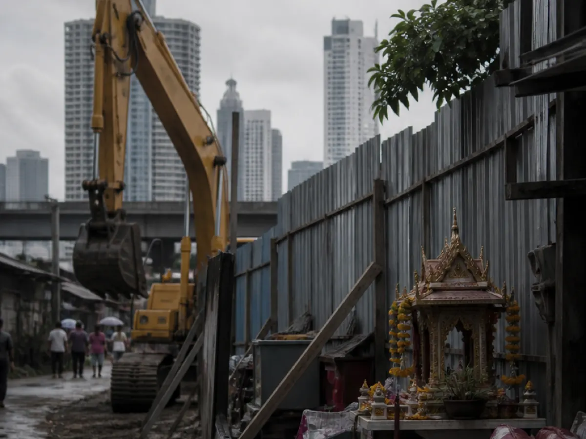 An excavator and a hoarding at the edge of a Bangkok community being cleared, a spirit house left standing