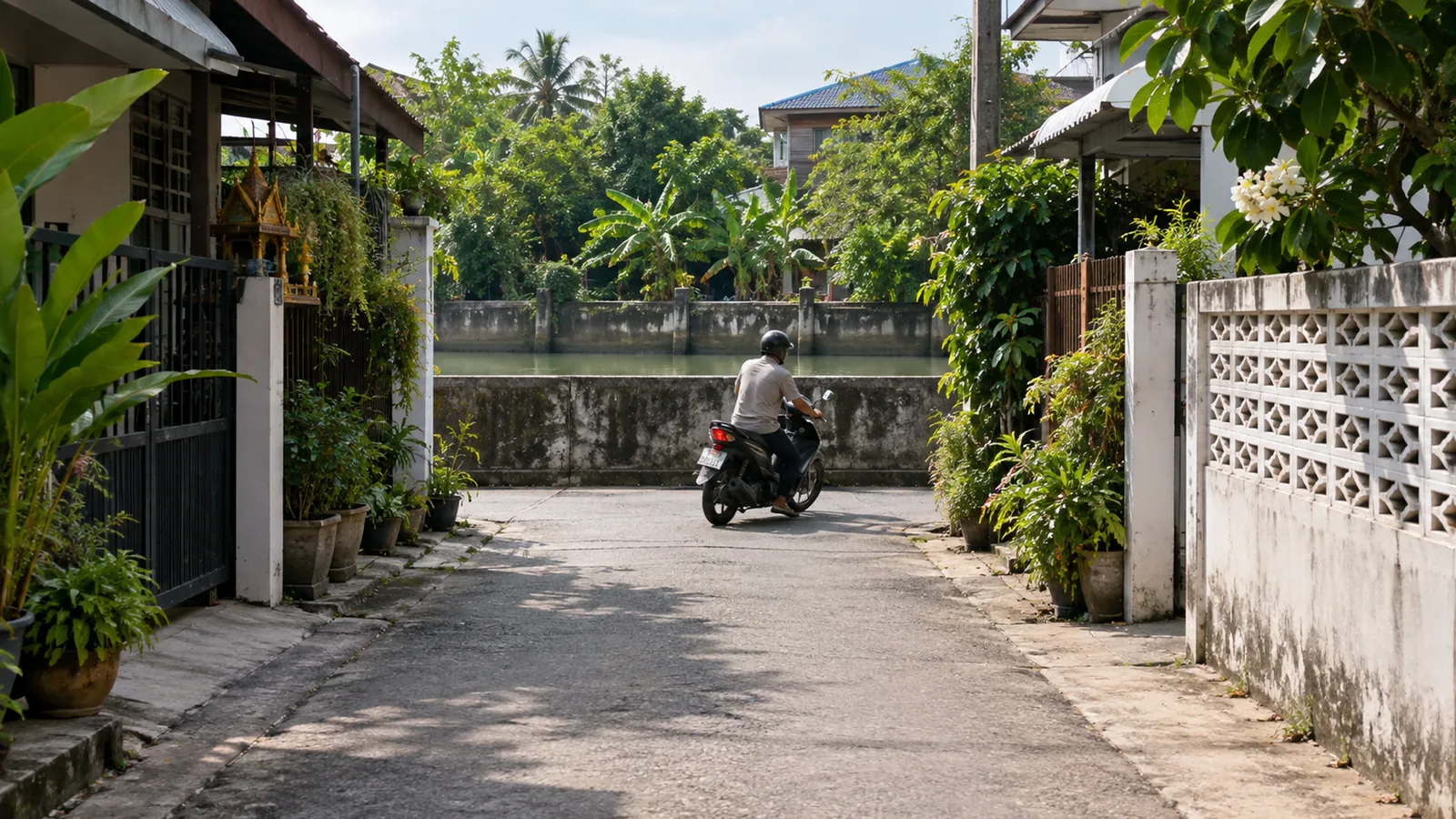 A narrow soi between garden walls ends at a concrete canal embankment, where a motorcyclist turns